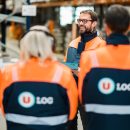 Quatre employés de U LOGISTIQUE en blouson orange et bleu, dont trois de dos et un de face souriant et portant des lunettes. Ils sont dans un entrepôt avec des racks en arrière-plan.