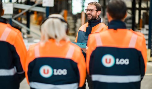 Quatre employés de U LOGISTIQUE en blouson orange et bleu, dont trois de dos et un de face souriant et portant des lunettes. Ils sont dans un entrepôt avec des racks en arrière-plan.