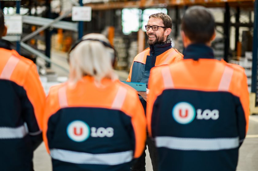 Quatre employés de U LOGISTIQUE en blouson orange et bleu, dont trois de dos et un de face souriant et portant des lunettes. Ils sont dans un entrepôt avec des racks en arrière-plan.