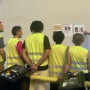 Un groupe de six employés de U LOGISTIQUE en gilets de sécurité jaunes et oranges, dont cinq de dos, écoutant un formateur qui pointe du doigt une affiche sur un mur.