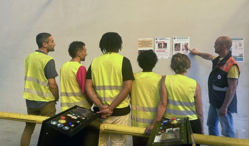 Un groupe de six employés de U LOGISTIQUE en gilets de sécurité jaunes et oranges, dont cinq de dos, écoutant un formateur qui pointe du doigt une affiche sur un mur.