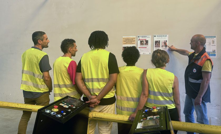 Un groupe de six employés de U LOGISTIQUE en gilets de sécurité jaunes et oranges, dont cinq de dos, écoutant un formateur qui pointe du doigt une affiche sur un mur.